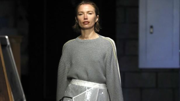 Eine Frau läuft in einem grauen Pullover, einem Paillettenrock und gelben Schuhen über einen Laufsteg.