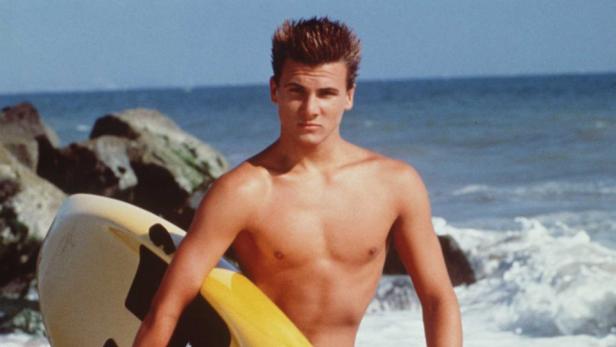 Ein junger Mann mit Surfbrett steht am Strand.