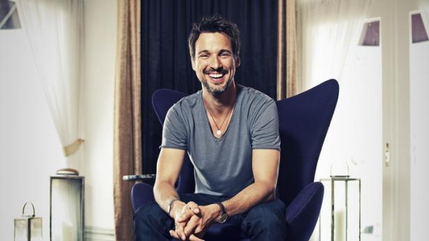 Ein lächelnder Mann sitzt in einem Sessel in einem eleganten Raum.