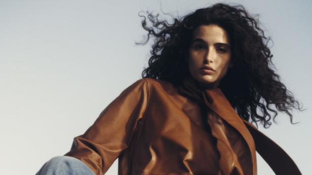 Eine Frau mit lockigem Haar posiert in Jeans, Lederbluse und Cowboystiefeln.