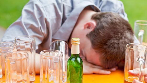Ein betrunkener Mann schläft mit dem Kopf auf dem Tisch, umgeben von leeren Gläsern und einer Flasche.