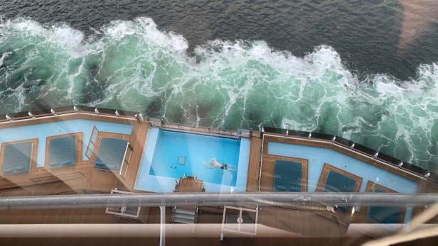 Eine Person schwimmt im Pool eines Kreuzfahrtschiffes, während Wellen daneben ans Ufer schlagen.