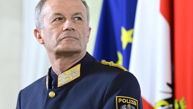 Ein älterer Mann in österreichischer Polizeiuniform steht vor EU- und österreichischer Flagge und blickt ernst zur Seite.