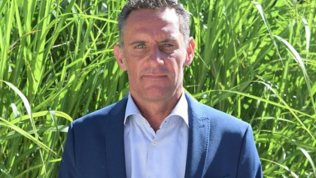 Ein Mann im blauen Anzug steht vor hohen grünen Gräsern und blickt ernst in die Kamera.