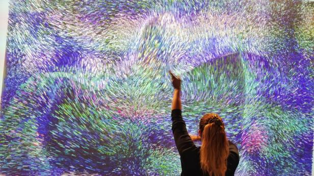 Eine Frau zeigt auf ein großes, abstraktes Kunstwerk mit blauen und grünen Farbtönen.