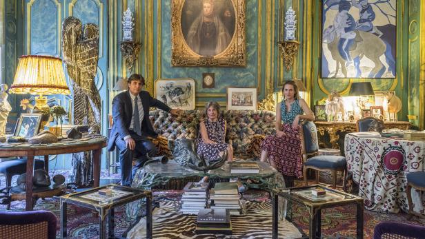 Drei Personen sitzen in einem opulenten Wohnzimmer mit Gemälden und Antiquitäten.