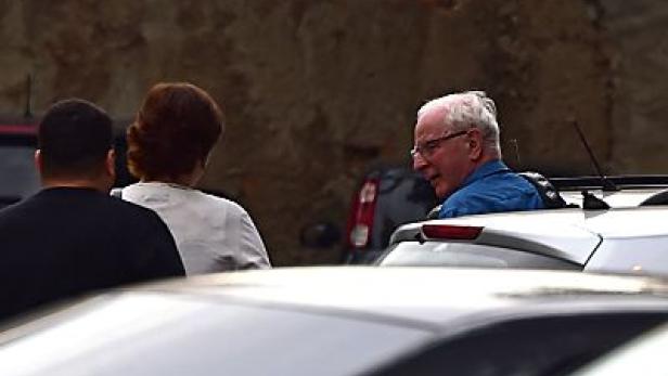 Ein älterer Mann mit Brille steht neben einem Auto, umgeben von anderen Personen.