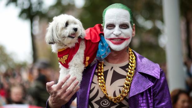 Ein Mann, als Joker verkleidet, trägt einen Hund im Wonder-Woman-Kostüm auf der Schulter.