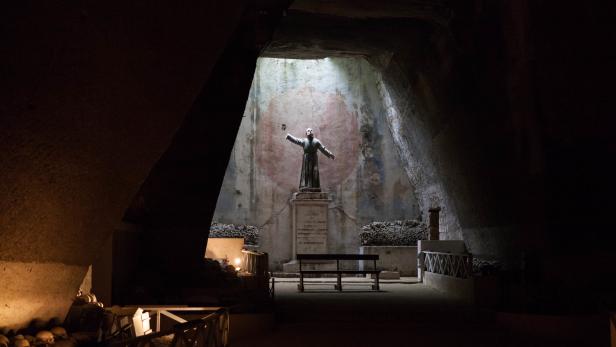 In einer dunklen Höhle steht eine Statue mit ausgebreiteten Armen auf einem Sockel.