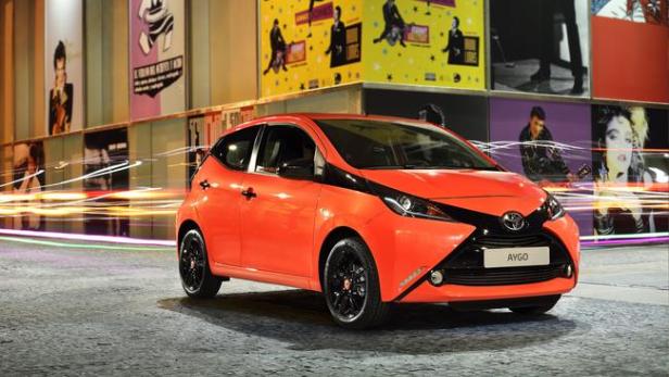 Ein orangefarbener Toyota Aygo vor einer Wand mit Musikplakaten.