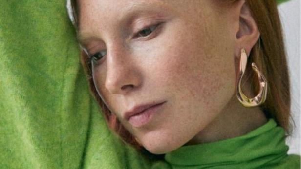 Eine Frau mit roten Haaren und Sommersprossen trägt einen grünen Rollkragenpullover und goldene Ohrringe.