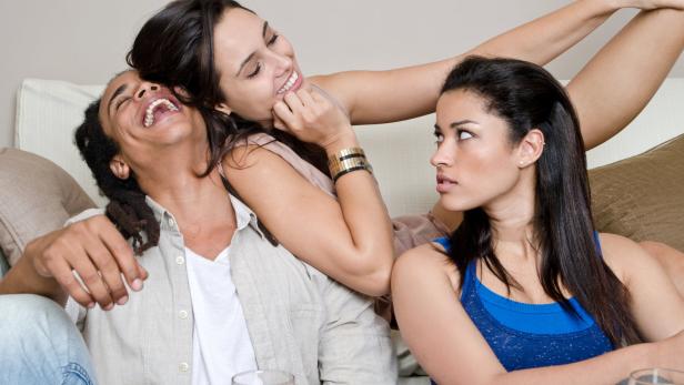 Drei junge Erwachsene sitzen zusammen auf einem Sofa, wobei eine Frau eifersüchtig wirkt, während die anderen beiden lachen.