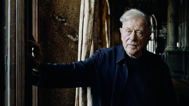 Ein älterer Mann in dunkler Kleidung steht an einem Fenster.