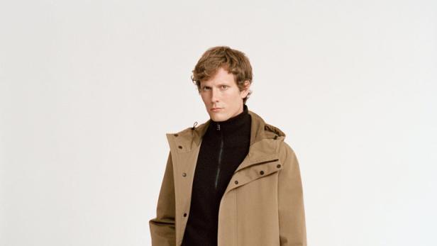 Ein Mann mit braunem Mantel, schwarzem Rollkragenpullover und schwarzen Stiefeln steht vor einem weißen Hintergrund.