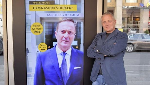 Ein Mann posiert vor einer digitalen Werbetafel, die für das Goethe-Gymnasium wirbt.