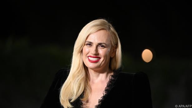 Rebel Wilson lächelt in einem schwarzen Kleid mit roten Lippen.