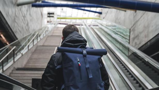 Ein Mann mit blauem Rucksack fährt eine Rolltreppe in einem modernen Gebäude hinauf.
