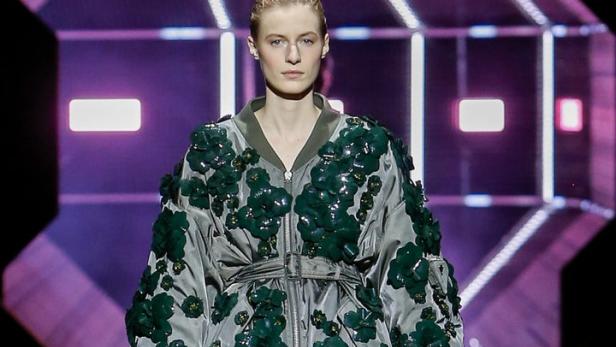 Ein Model auf dem Laufsteg trägt eine graue Bomberjacke mit grünen Blumenapplikationen und einen goldenen Rock.