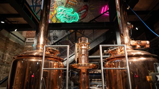 Zwei große, glänzende Kupferkessel in einer Brauerei, im Hintergrund Wandmalereien und Neonlichter.