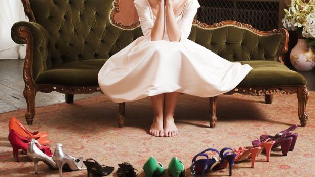 Eine Frau im Kleid sitzt auf einem Sofa, umgeben von vielen Schuhen.