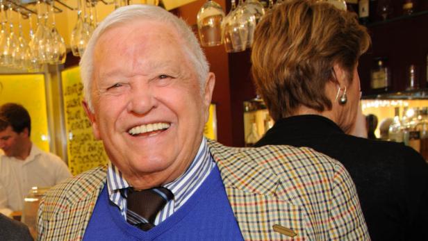 Ein lächelnder älterer Mann mit kariertem Sakko steht in einem Restaurant.