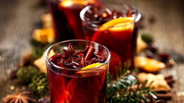Drei Gläser Glühwein, garniert mit Sternanis, Zimtstangen und Orangenscheiben, auf einem Holztisch.