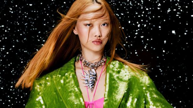 Eine Frau mit orangefarbenem Haar trägt einen grünen Paillettenanzug und eine auffällige Halskette vor einem dunklen Hintergrund.