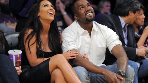 Kim Kardashian und Kanye West schauen bei einem Basketballspiel lachend nach oben.