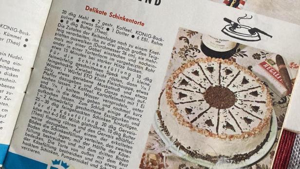 Eine Schinkentorte, eine Flasche Wein und eine Zigarrenschachtel auf einer Tischdecke.