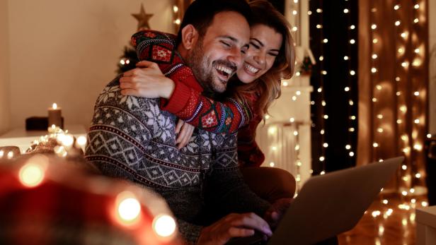 Ein lächelndes Paar mit Weihnachtspullovern sitzt vor einem Laptop, umgeben von Lichtern.