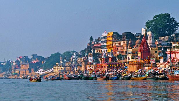 Varanasi_ cc by J Kaps.jpg Boote liegen am Ufer des Ganges in Varanasi, Indien, vor einer bunten Stadtkulisse.