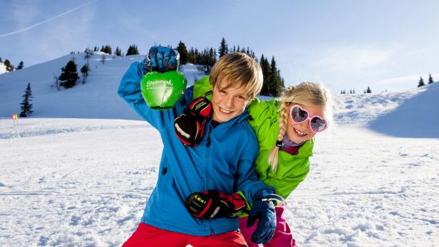 Zwei Kinder haben Spaß im Schnee, eines hält einen grünen Herzanhänger der Steiermark hoch.