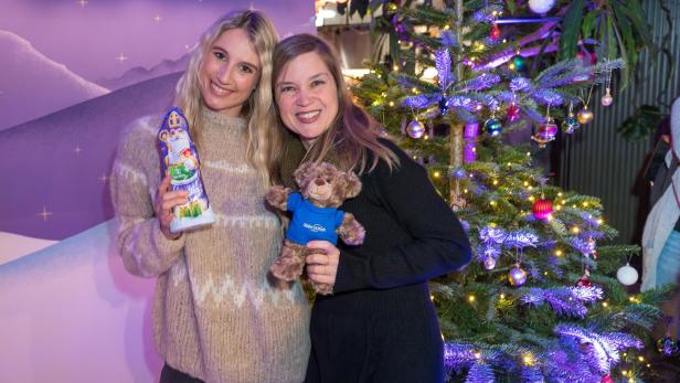 Zwei Frauen posieren mit einem Schokoladen-Nikolaus und einem Teddybär vor einem Weihnachtsbaum.