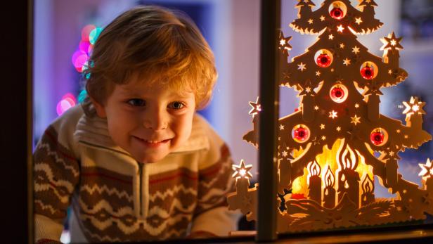 Ein lächelnder Junge schaut durch das Fenster auf einen hölzernen Weihnachtsbaum.
