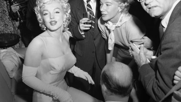 Marilyn Monroe, umgeben von Menschen, sitzt auf einem Sofa.