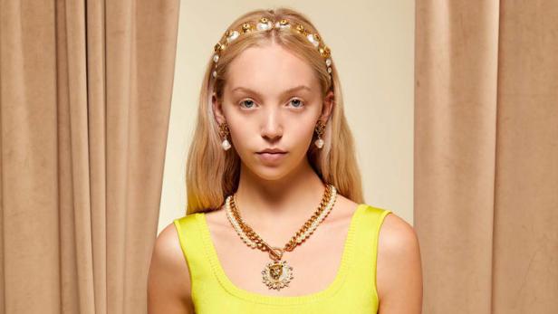 Eine blonde Frau trägt ein gelbes Kleid, Schmuck und ein Haarband vor einem beigen Hintergrund.