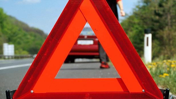 Ein rotes Warndreieck steht auf einer Landstraße vor einem defekten Auto.