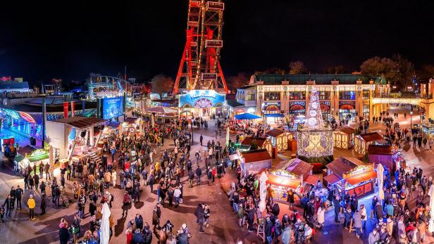 Der Wiener Prater mit dem beleuchteten Riesenrad und vielen Menschen bei Nacht.