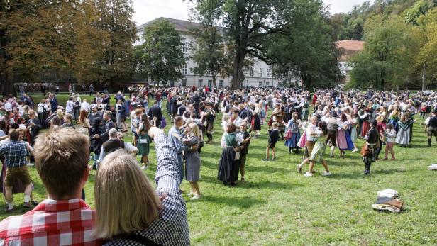 Eine große Menschenmenge in Tracht tanzt auf einer Wiese vor einem Gebäude.