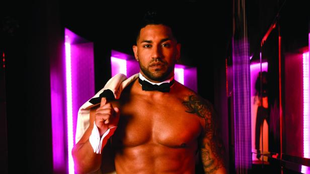 Ein muskulöser Mann mit Tätowierungen, Fliege und offenem Jackett steht in einem pinkfarbenen Gang.