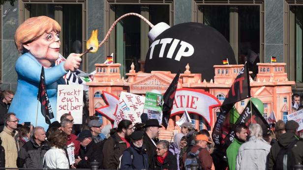 Eine Anti-TTIP-Demonstration mit einer Merkel-Figur, die eine Bombe mit der Aufschrift „TTIP“ anzündet.