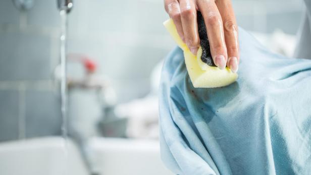 Eine Hand reinigt einen Fleck auf einem hellblauen Kleidungsstück mit einem Schwamm.