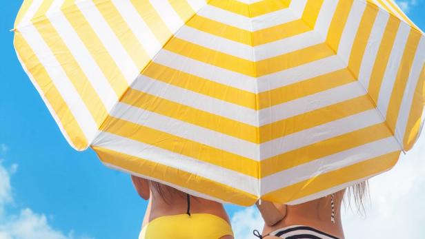 Zwei Frauen stehen mit dem Rücken zur Kamera unter einem gelb-weiß gestreiften Sonnenschirm.