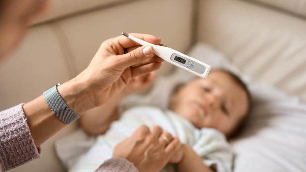 Eine Hand hält ein Fieberthermometer mit 38,1°C, während ein kleines Kind im Hintergrund liegt.