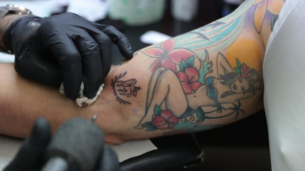 Ein Tätowierer sticht ein neues Tattoo auf den Arm einer Person mit bestehenden Tätowierungen.