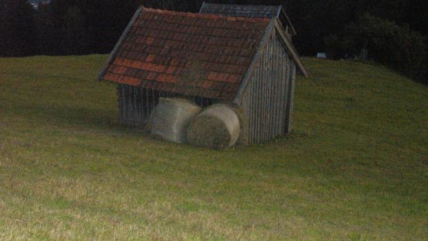 Ein kleines Holzhaus mit zwei Heuballen davor auf einer Wiese.