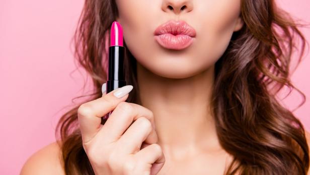 Eine Frau hält einen pinkfarbenen Lippenstift vor ihren geschminkten Lippen.