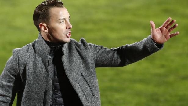 Ein Mann in einem grauen Mantel gestikuliert mit erhobener Hand.