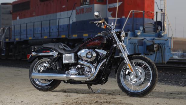 Eine schwarze Harley-Davidson Low Rider vor einem roten und blauen Zugwaggon.