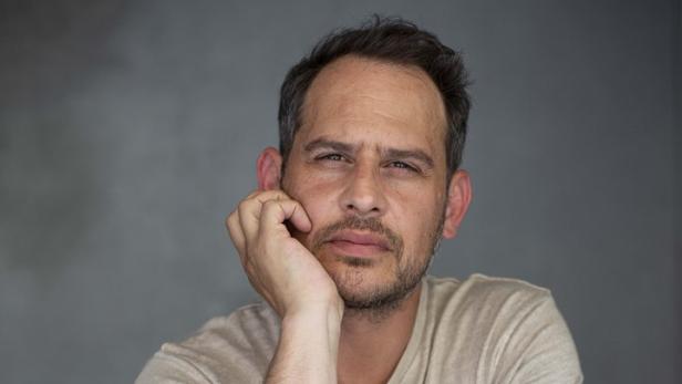 Ein Mann mit Tattoos sitzt auf einem Stuhl und stützt seinen Kopf in die Hand.